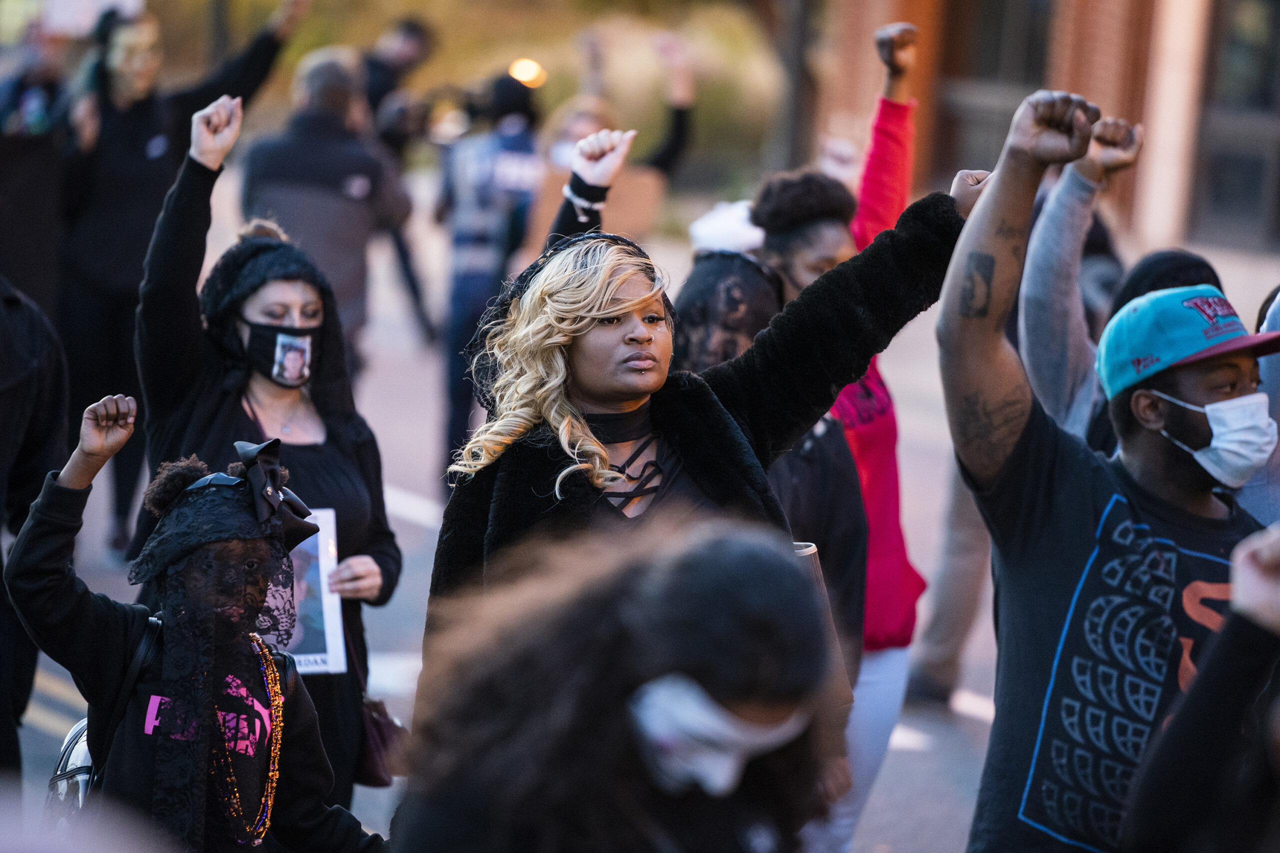 A group of protesters stand with their left or right hand up in a fist. Almost everyone wears a face mask.