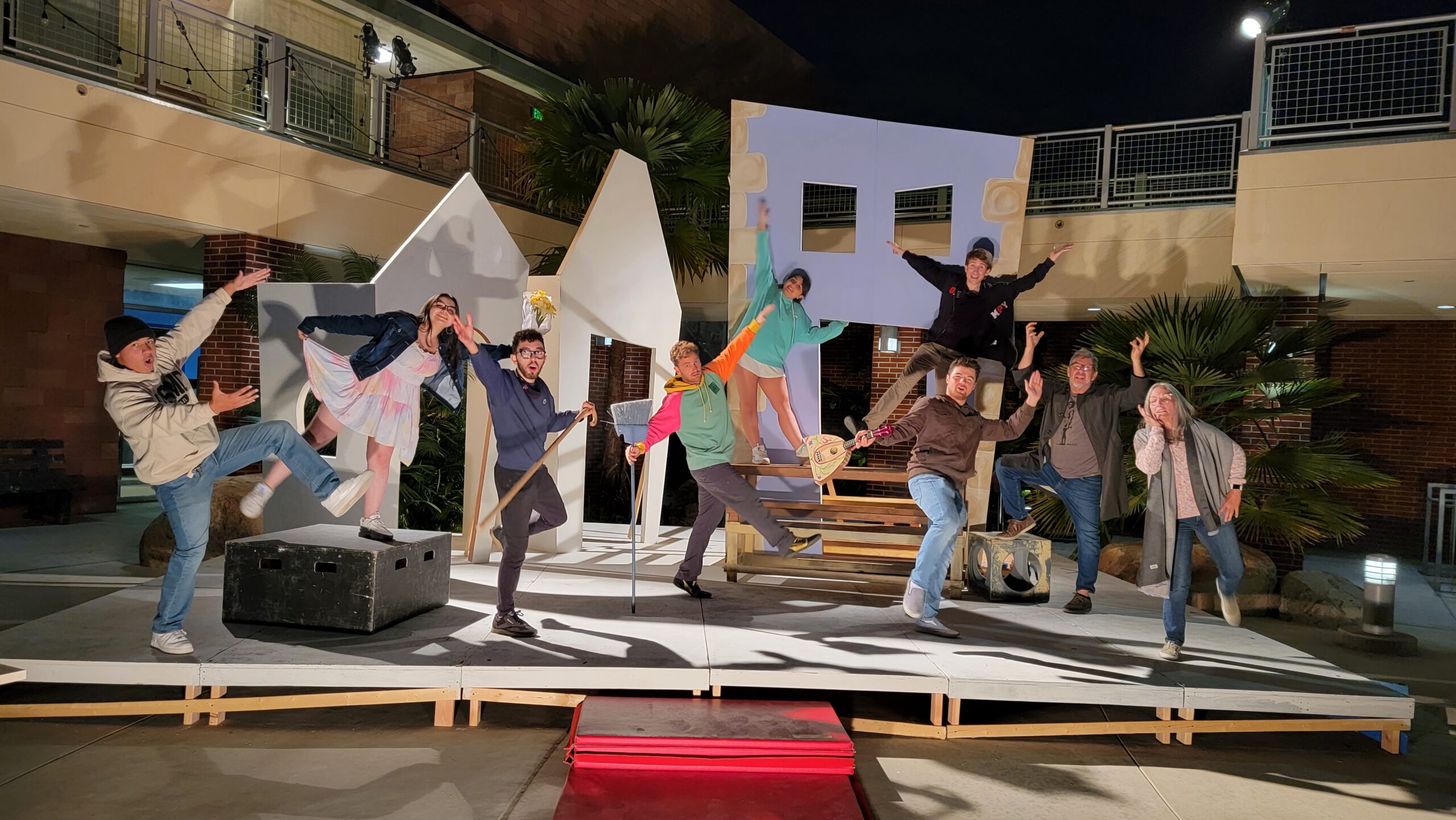 A group of actors pose on stage at a small plaza with a second-story building behind them on the left and an overhead walkway in the middle and right.