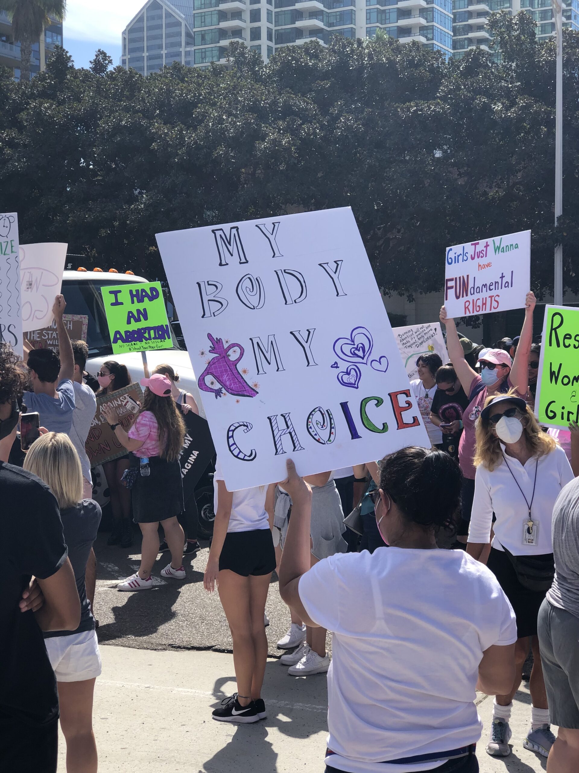A group of women protests and carries signs in a park in downtown San Diego. The woman in front carries a sign that says, "MY BODY MY CHOICE."