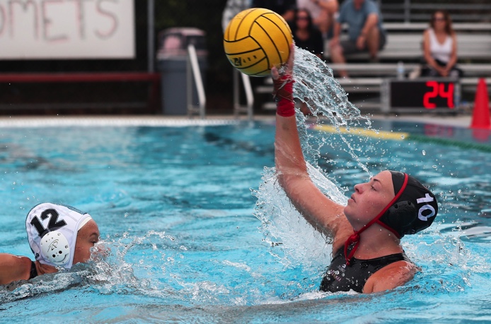 A female water polo player on the right throws a yellow ball with her right hand as an opponent swims toward her.