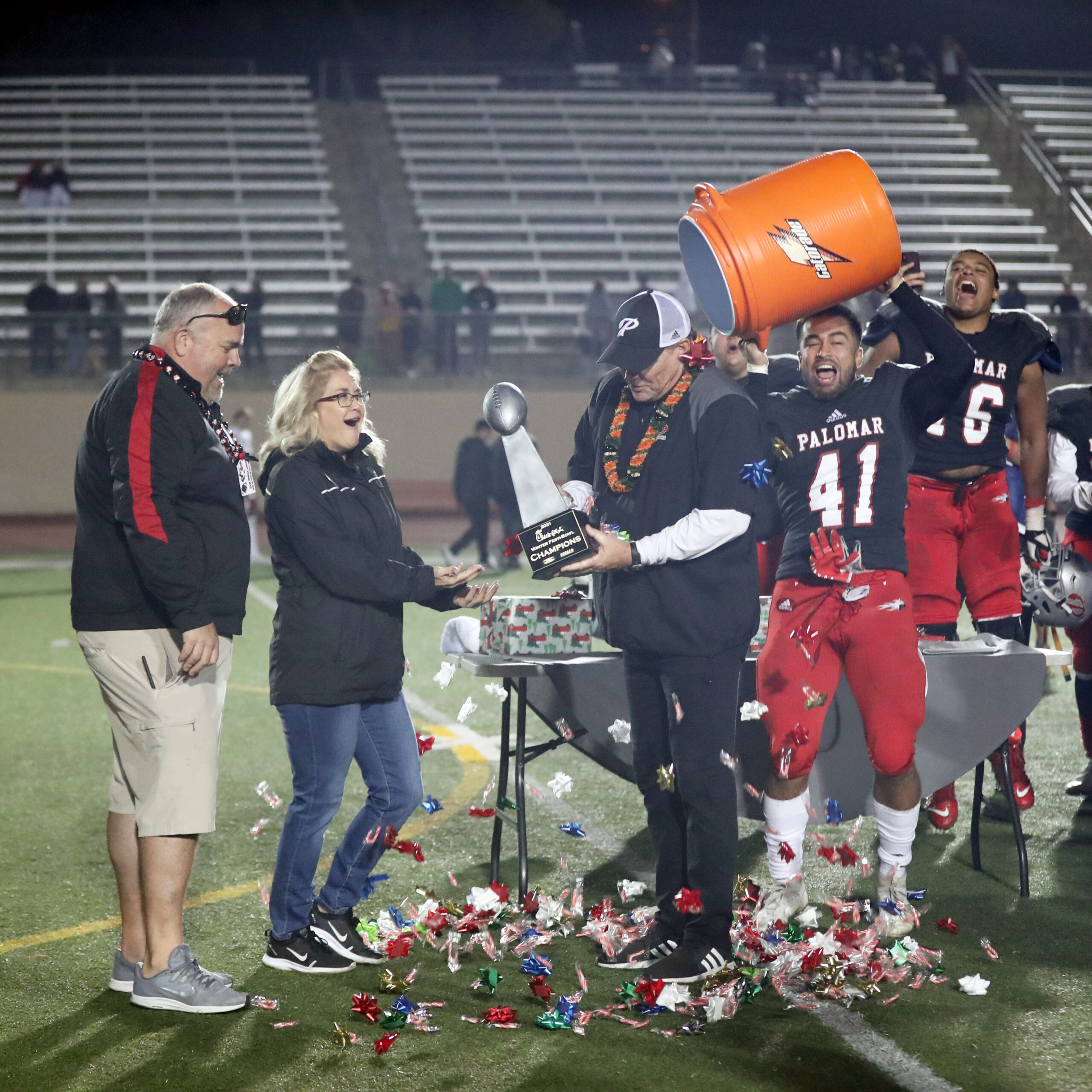 A football coach holds a trophy as a football player dumps ribbons and candy from a large Gatorade bin. Several people around them laugh.