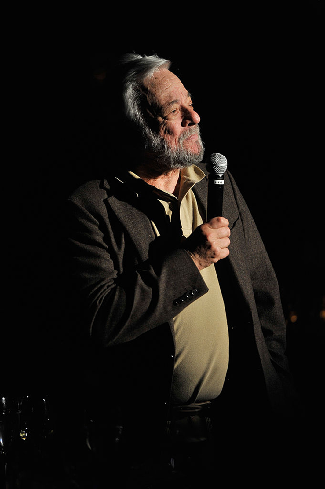Composer Stephen Sondheim speaks with a microphone in his right hand. A spotlight shines on him, casting a sharp shadow contrast of the front of his body and his face with the black background.