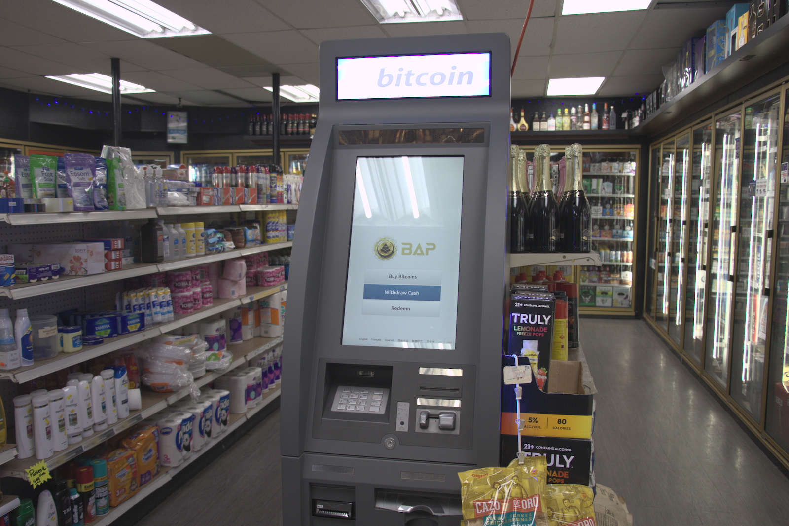 An ATM that accepts Bitcoin stands inside a liquor store by toiletries and drinks in a fridge.