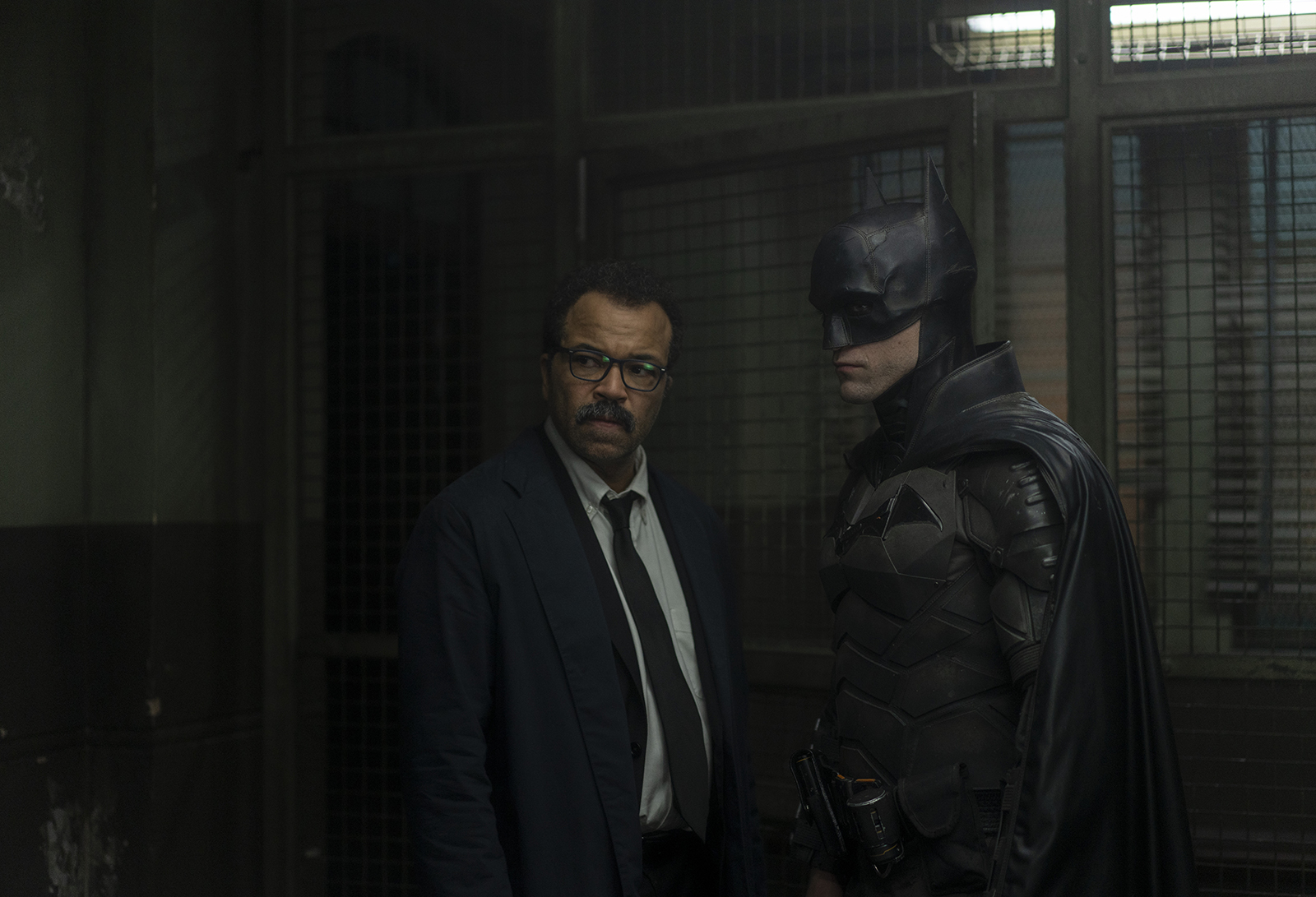 Commissioner James Gordon talks to Batman as they gaze to the right in a police station.