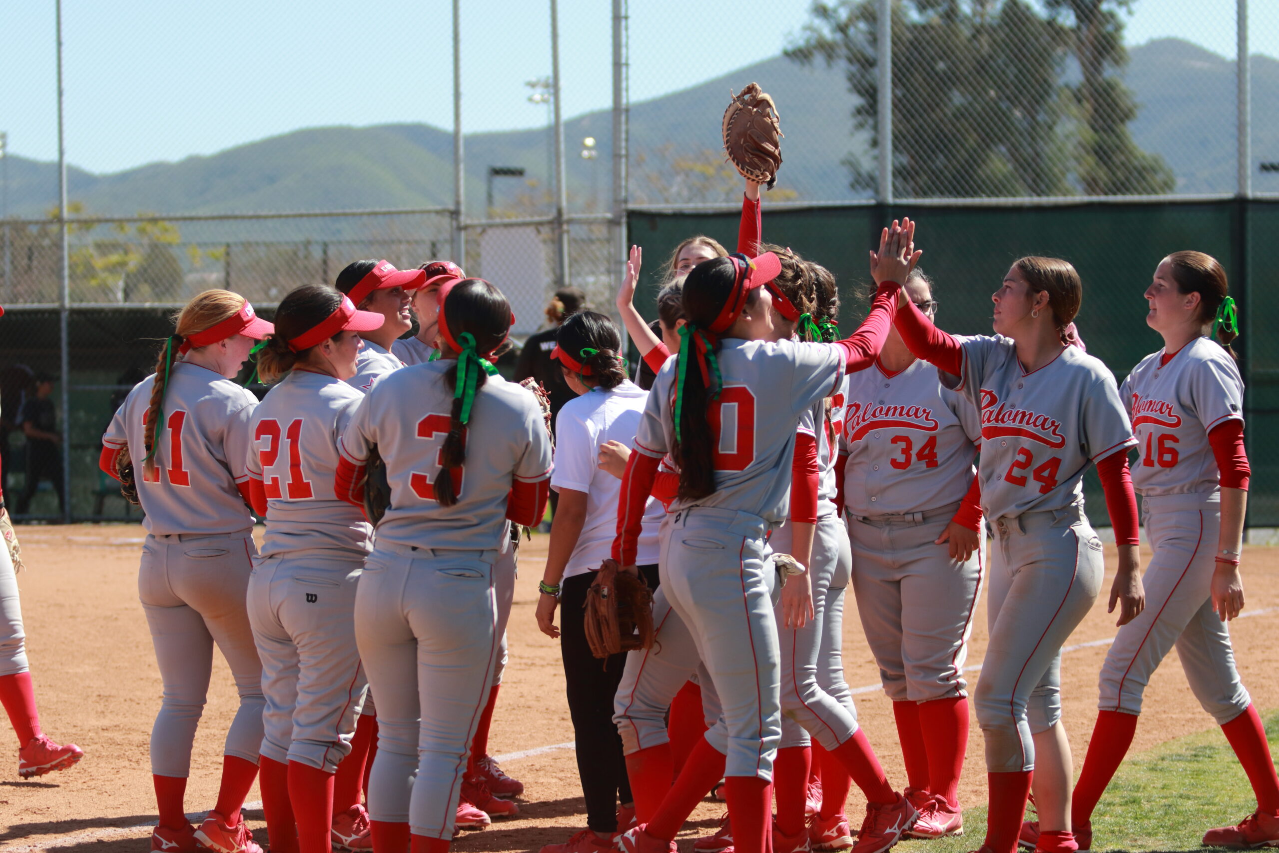 A group of female softball players gather around each other after a game in a field. Two of them give each other a high-five.