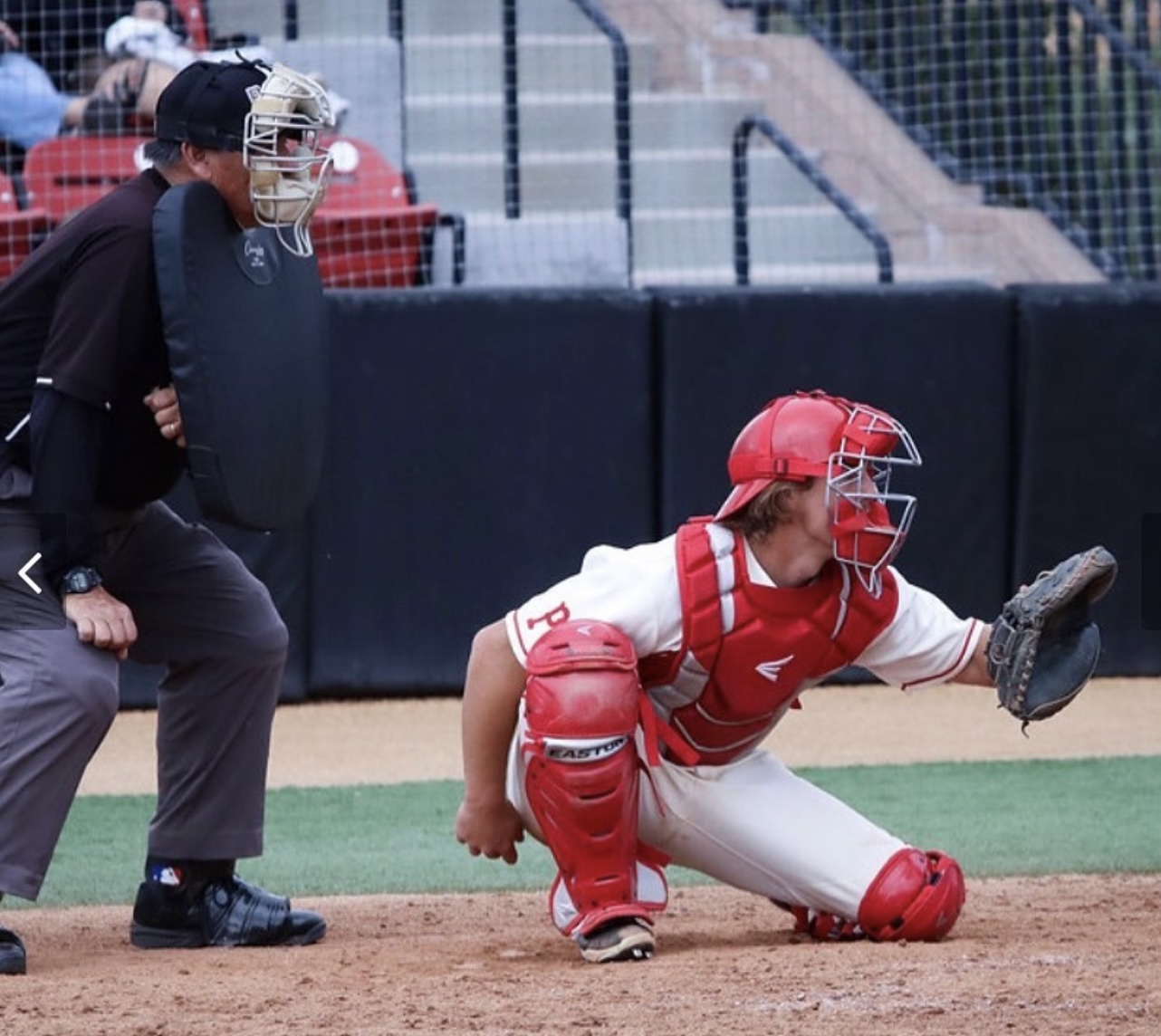 A male Palomar catcher kneels and prepares to catch a baseball with his mitted left hand while an umpire half-squats behind him.