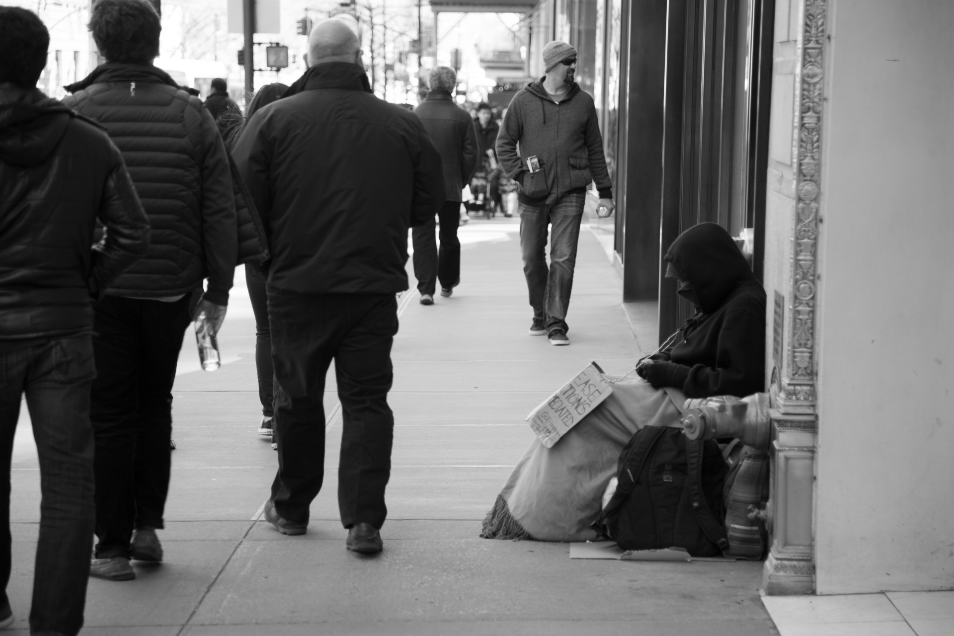 Homeless sitting on a street with sign with people walking by him.