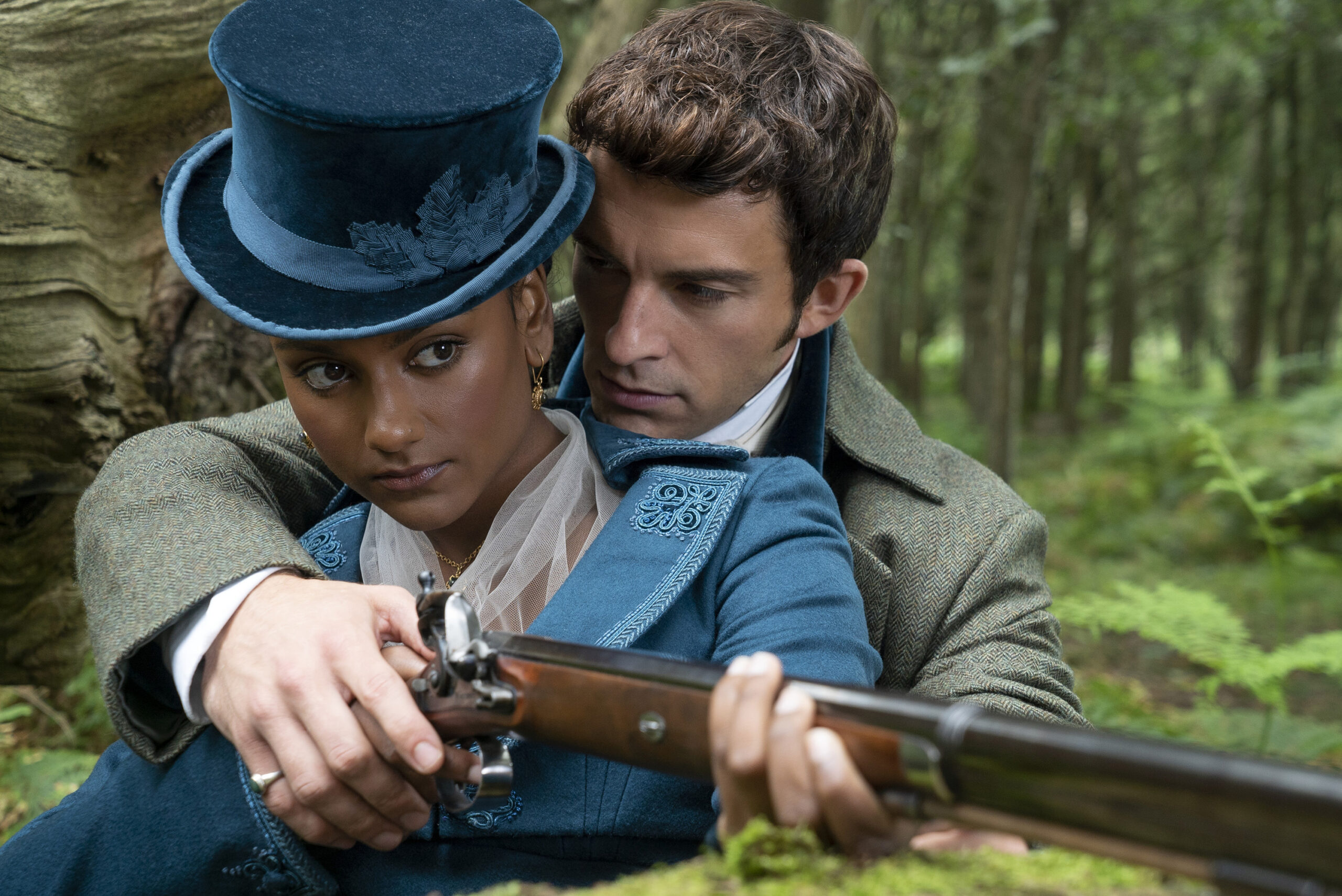 A woman holds and aims a musket rifle while a man holds her hand and rifle with his face close to her neck. She wears a blue top hat and coat, and he wears a grey jacket.
