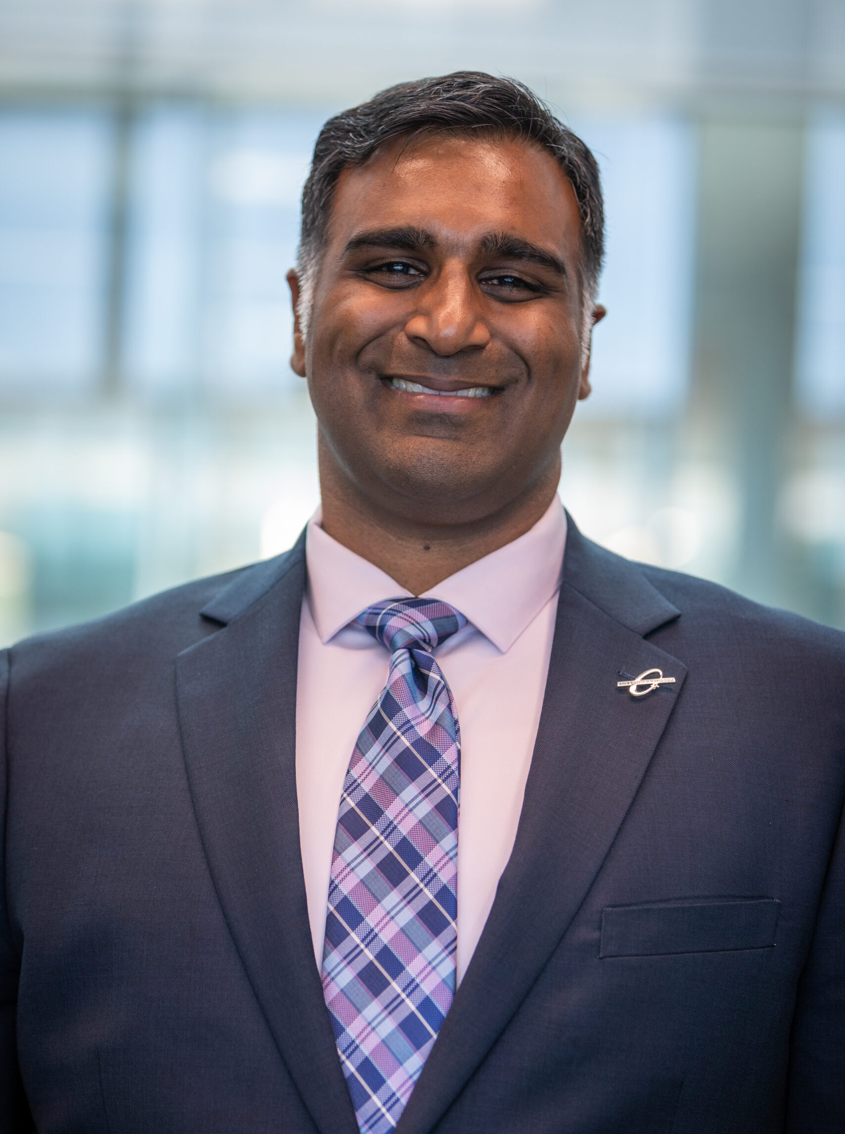 Portrait of a man of Indian descent smiling, wearing a dark grey suit and a checkered tie.