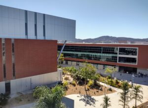 The Palomar library's brick and metal building looms over its neighboring Humanities building, which has a wall of glass windows. There are trees and sidewalks in between.