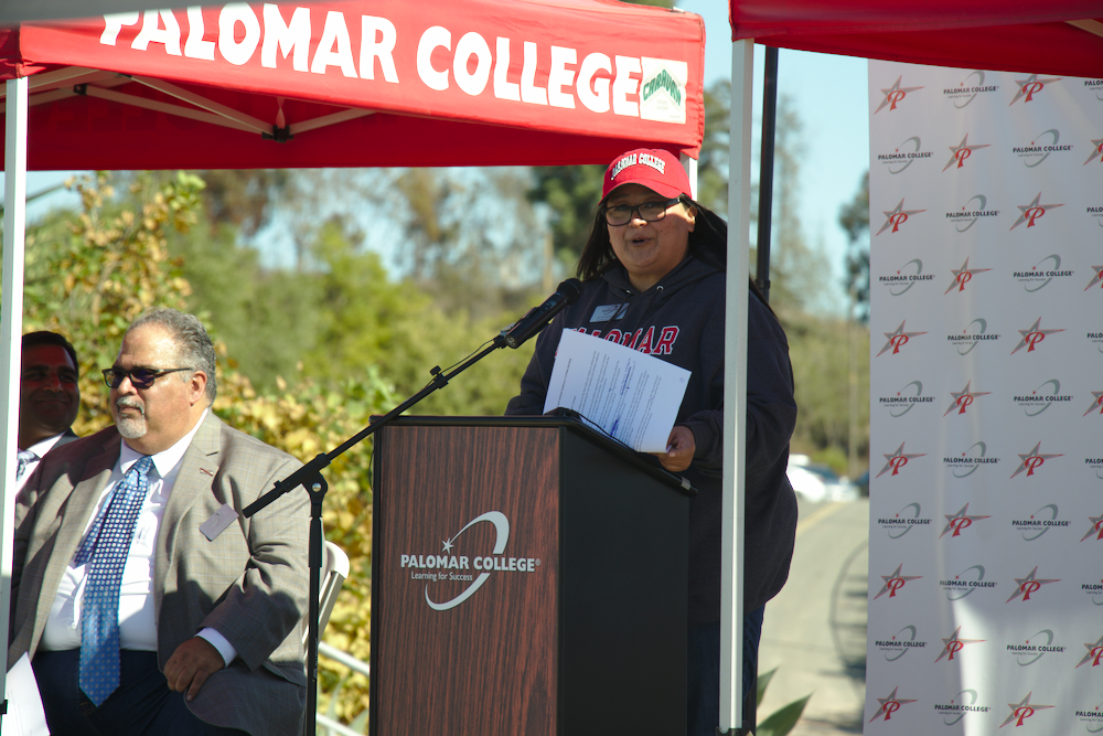 A woman wearing a black hoddie and a red cap speaks at a lectern.