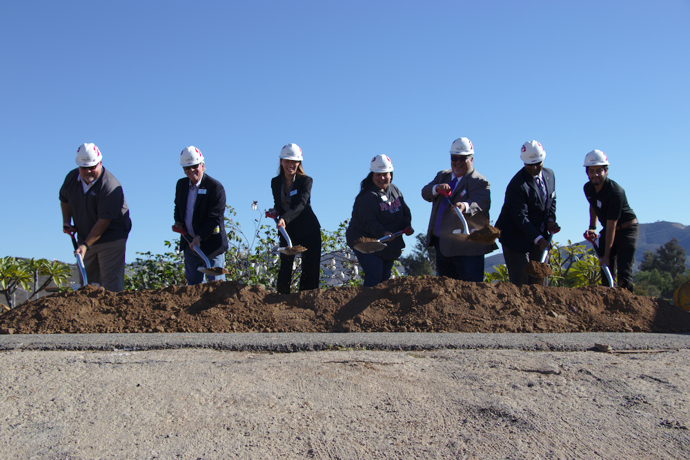 Seven people wearing white hard hats shovel dirt on a sunny day.