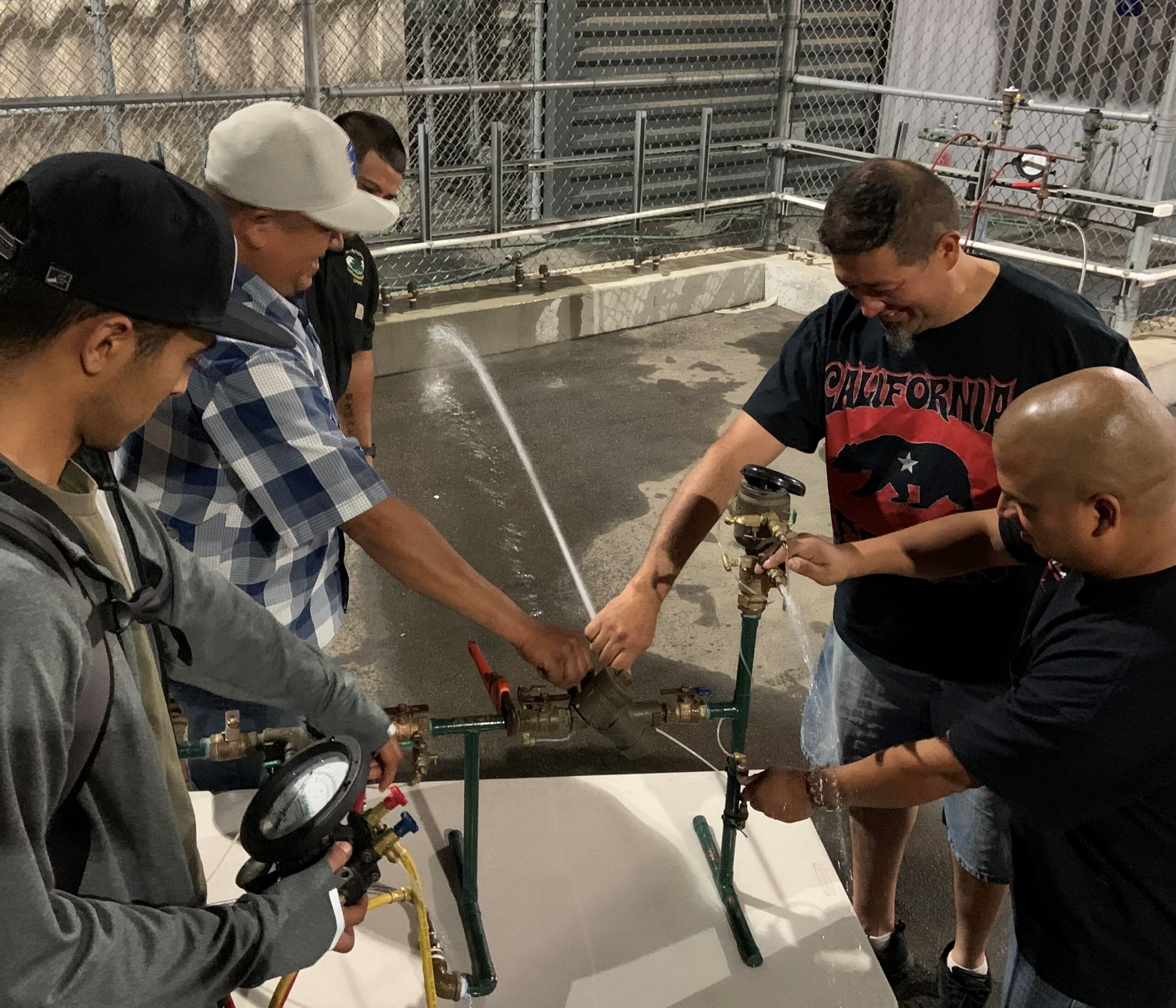 Five men gather around a water backflow device and tests it. The device sprays a strong stream of water at a concrete ledge.