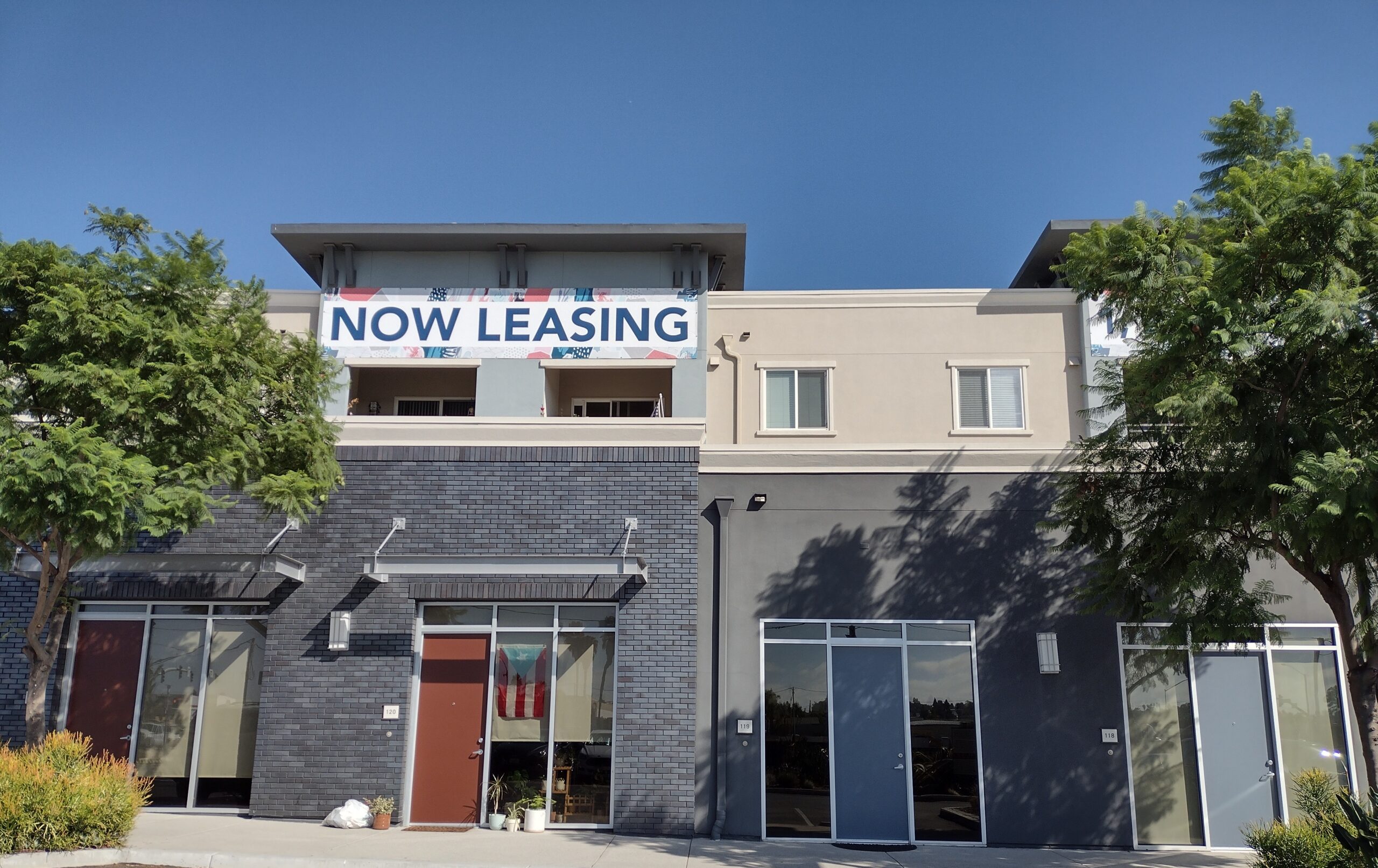 A Now Leasing sign is hung on the side of an apartment complex on Las Posas Avenue in San Marcos. The building is gray and beige with bricks and stucco facade.