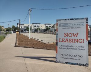 The photo has a sandwich board sign on a sidewalk that reads the Palomar Station Apartment Community has apartments for rent. The building itself can be seen in the background.