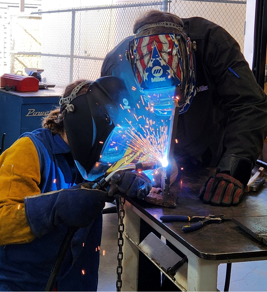 Two people weld a piece of metal, which gives off a blue light.