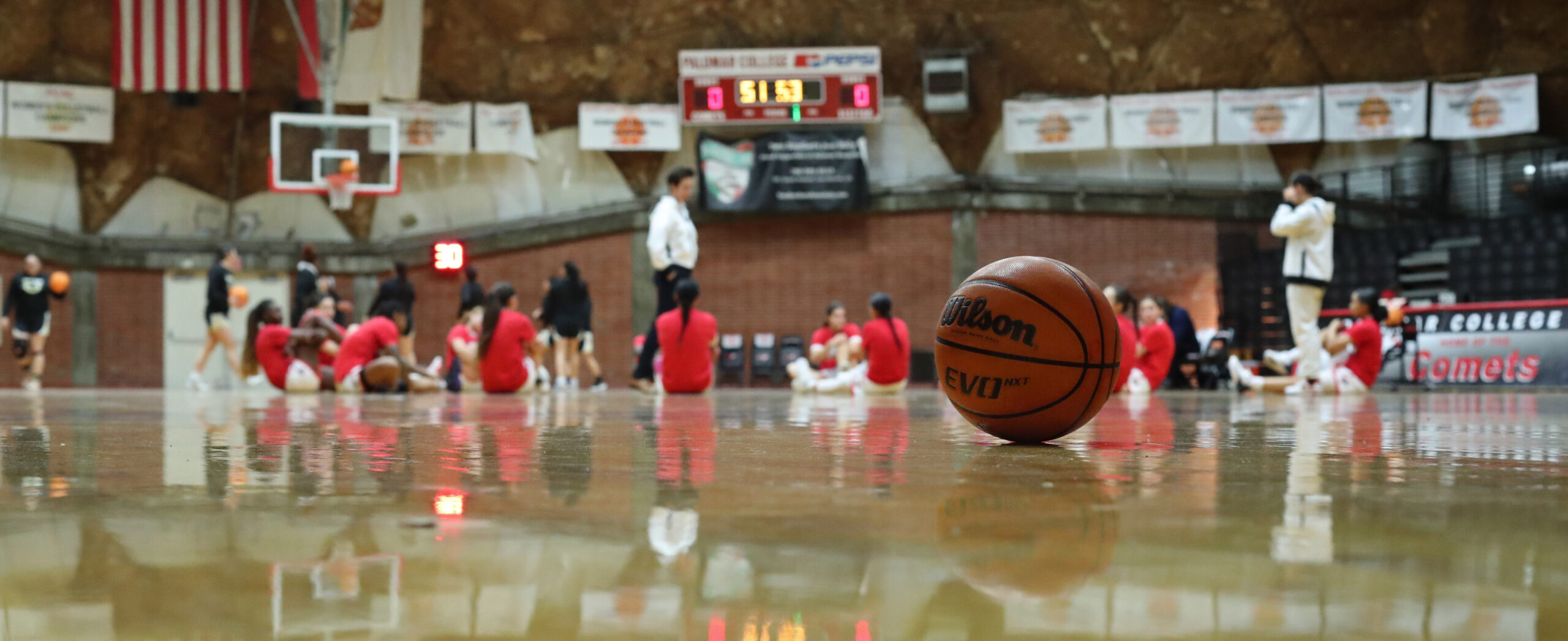 A basketball stands at a basketball court at Palomar College's Dome with the women's basketball team sit on the floor in a circle while listening to their coach.