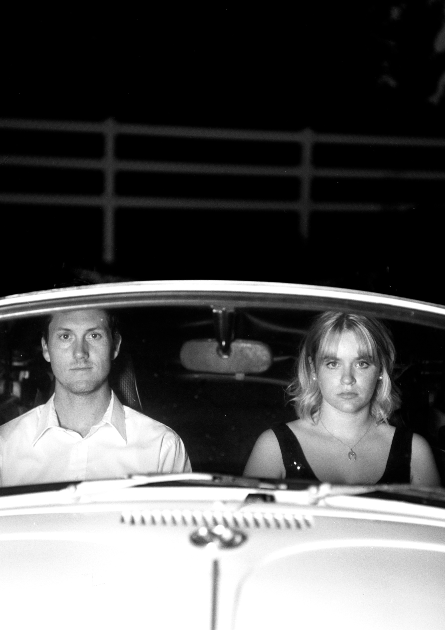 A man and woman are sitting in a convertible, facing the viewer. Black and white photo.