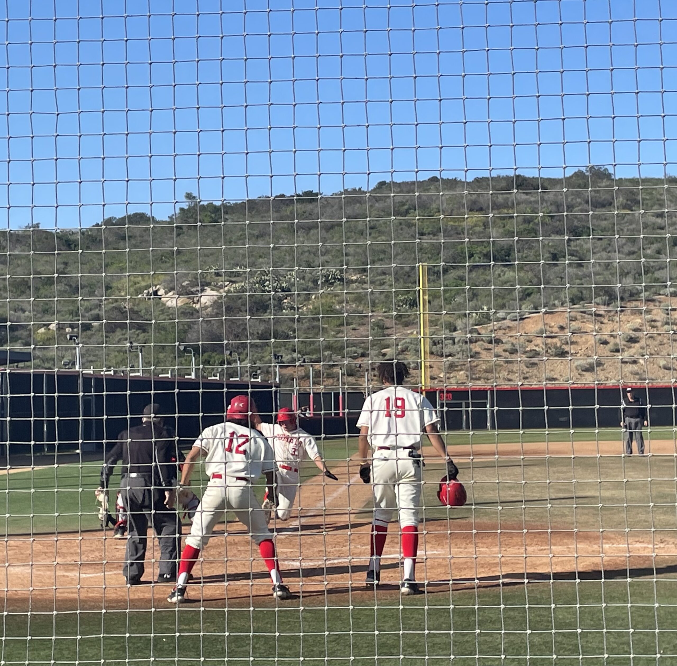 Two Palomar baseball players and an umpire watches another teammate slide into base. A net is in the foreground.