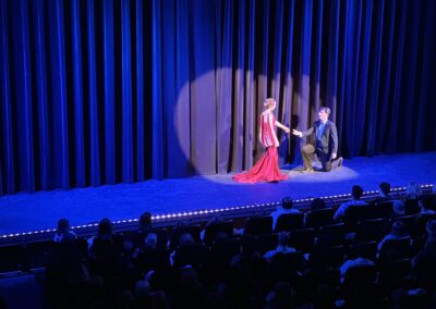 suited man kneels before a woman in red dress, on stage