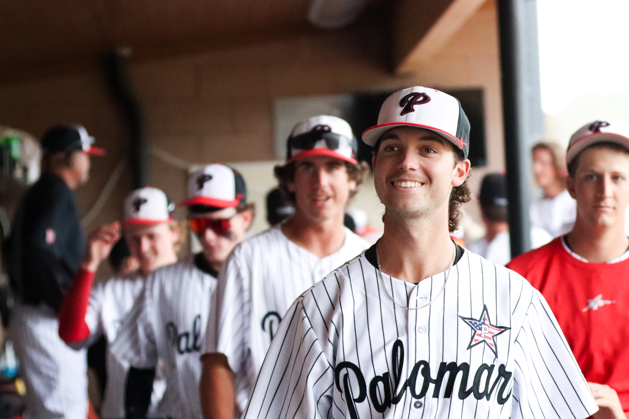 A male Palomar baseball player smiles to the photographer's left at the dugout with two fellow teammates look on from behind him. Several players and a coach are also behind them and are blurry.