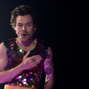 Harry Styles at a concert wearing sequin and putting his hand on his chest to thank his fans.