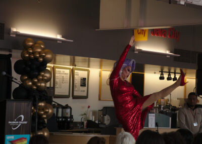 drag queen in a glittery red dress and purple wig kicks a leg in the air in front of a wall of rainbow balloons at a college event