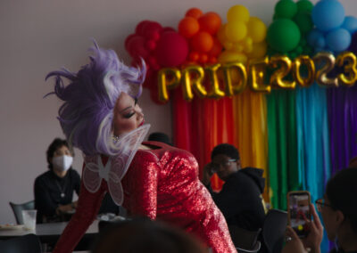 drag queen in a glittery red dress and purple wig performing expressively in front of a wall of rainbow balloons at a college event