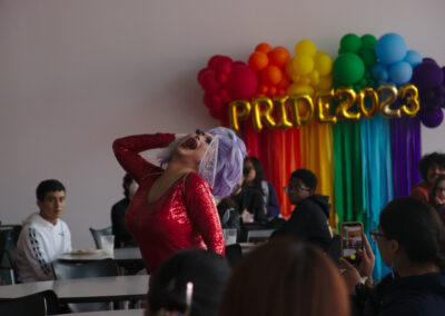 drag queen in a glittery red dress and purple wig performing expressively in front of a wall of rainbow balloons at a college event