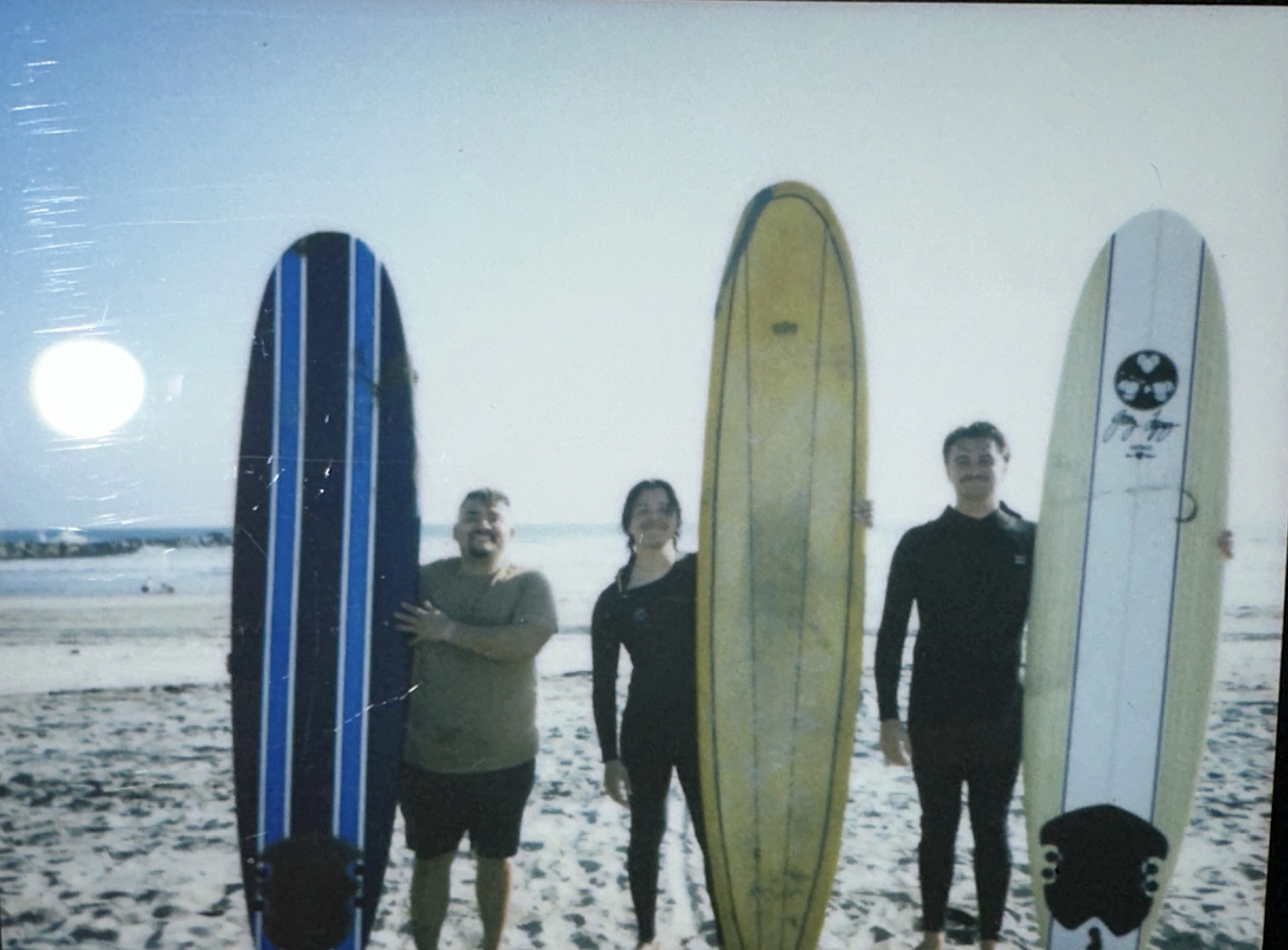 Three young men stand side by side and hold their surfboard upright at a beach on a sunny day.