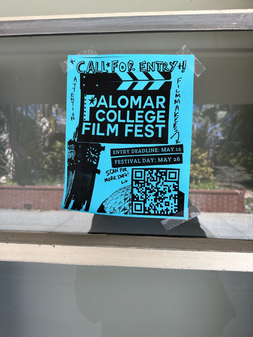 A teal-color paper flyer taped on a glass pane, promoting the 2023 Palomar College Film Fest on May 26 and encouraging students to submit their entry by May 12.