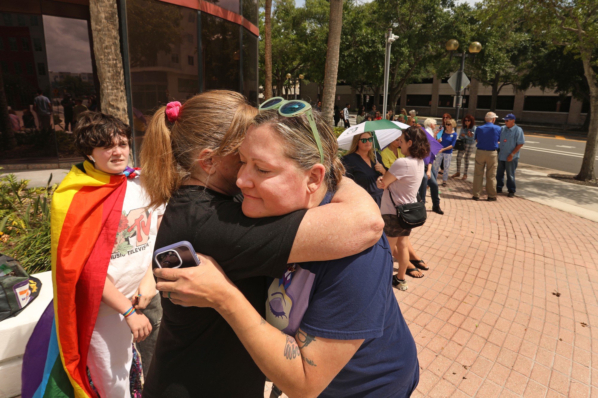 Two women hug each other on a sunny day. A young person stands to the left of them wearing a rainbow cloak. More people stand in the background on a large sidewalk by a glass office building.