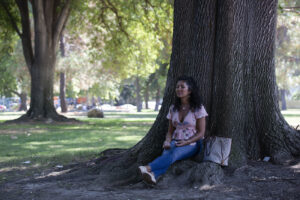 A woman sits beneath a large tree in a park with her legs straight and crossed in front of her. Her eyes are closed as if meditating. Her purse/bag is to her left.