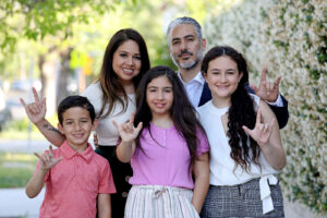 A family of five including a mother, father, two pre-teen daughters, and a young son stand close to each other, gesturing with their hand in sign language "I love you" where the thumb, index finger, and pinkie finger are pointed up.
