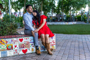 A young Hispanic couple sit together holding in each others' arms. Hearts and other icons are painting on the tiles that make up the planter that they're sitting.