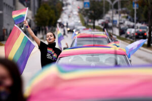A young woman waves a Pride flag in her right hand next to row of cars with a Pride flag draped on top of the car.