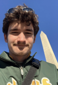 A man with curly brown hair looks down at the camera. He is standing in front of the Washington Monument.
