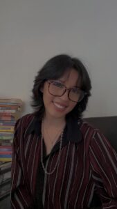 A brunette with large tortoise shell glasses cants her head and smiles. Behind her is a stack of books.