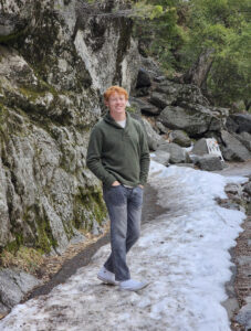 Red haired male stands in the snow outside. He wears a green sweater and jeans. He is smiling. 
