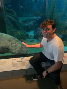 A man in a white shirt and black pants kneels in front of a glass aquarium with his hand outstretched like he is petting the large fis in the background.