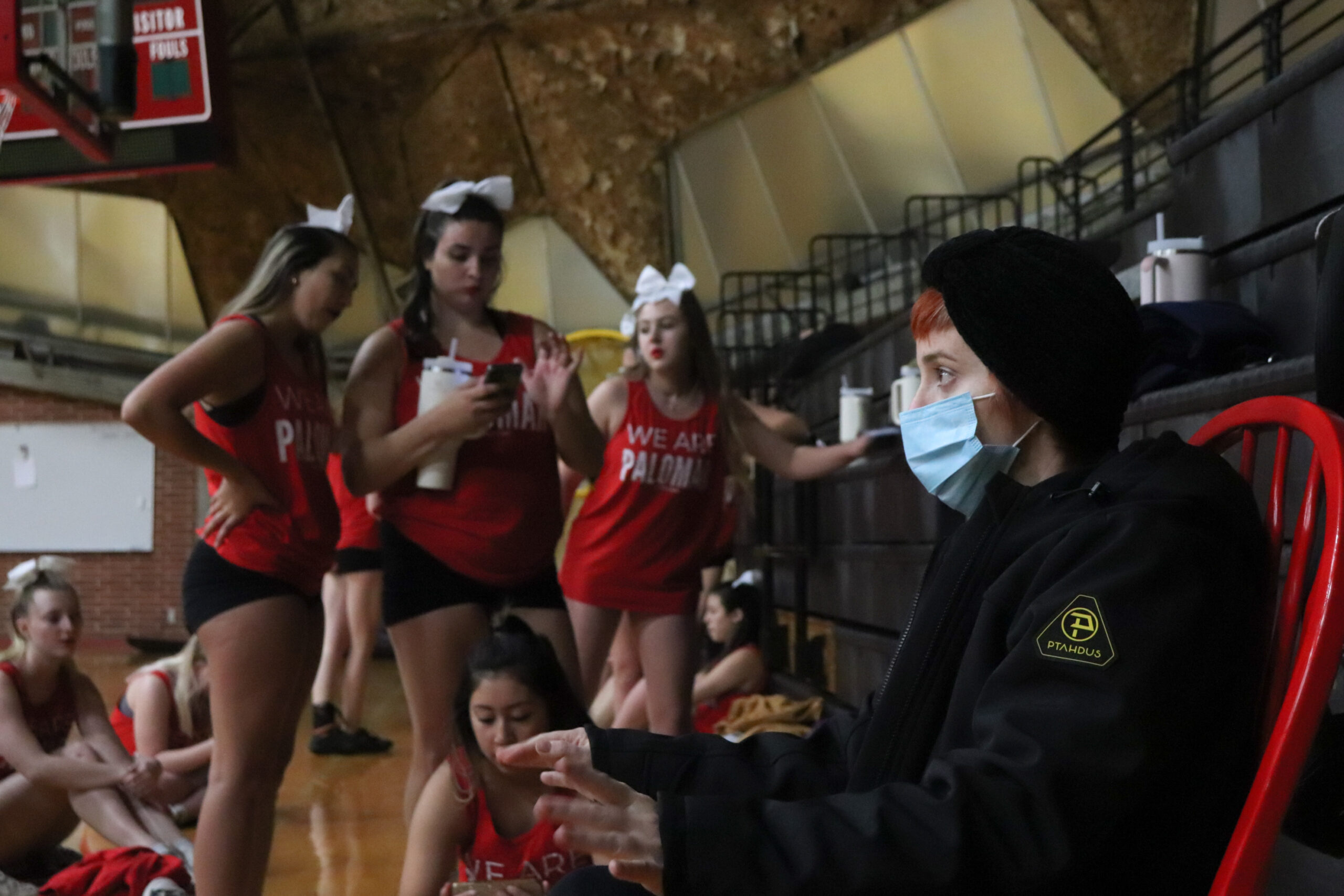 Coach Reed sits in her custom red rocking chair wearing a beanie and a face mask during a cheer practice.