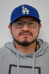 Headshot of a man with a blue baseball cap and glasses wearing a grey hoodie.