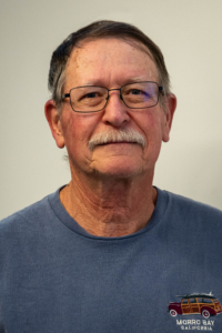 Headshot of a man with gray hair and glasses. He has a mustache and a blue shirt.
