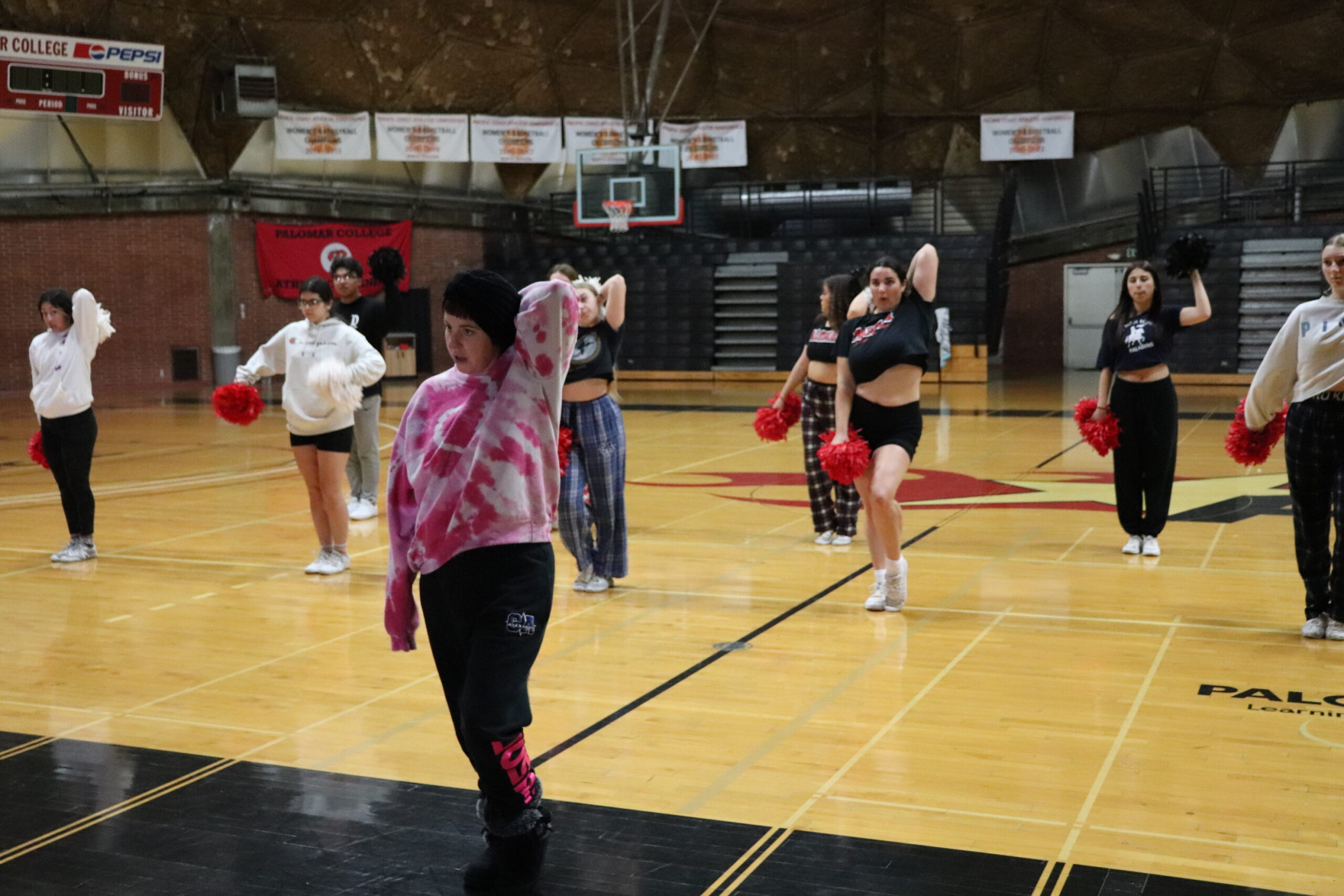 The Palomar Cheer coach, Coach Reed, stands in front of her team, showing them how to perform a new routine inside the Dome on the Palomar San Marcos campus. She's wearing a black beanie, pink tie dye seater, and black sweatpants.