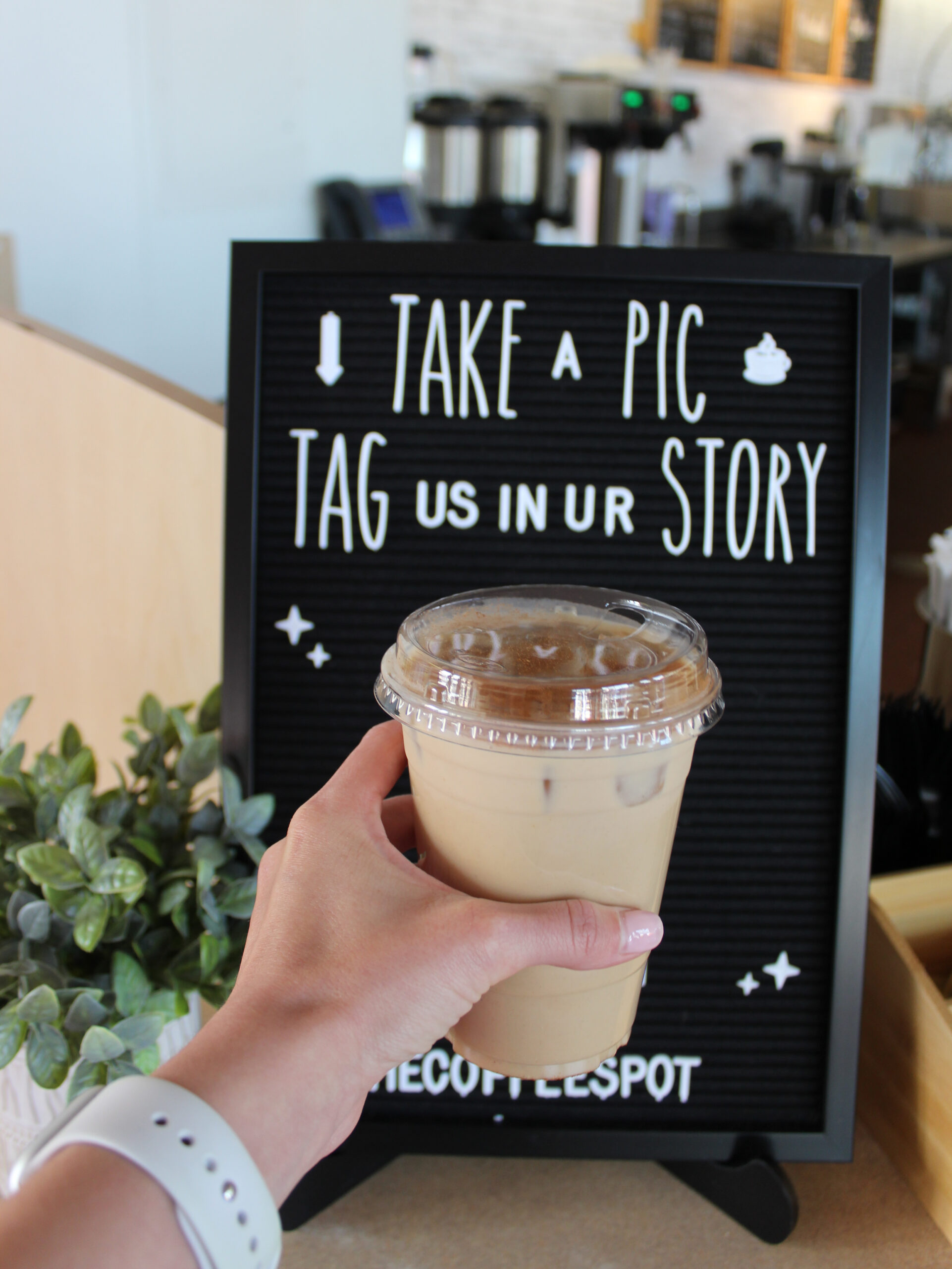 Hand holding a brown drink in front of a sign that reads "take a pic and tag us in your story"
