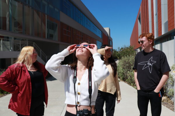 A group of college students stand outside while wearing solar eclipse glasses to look at the sun.