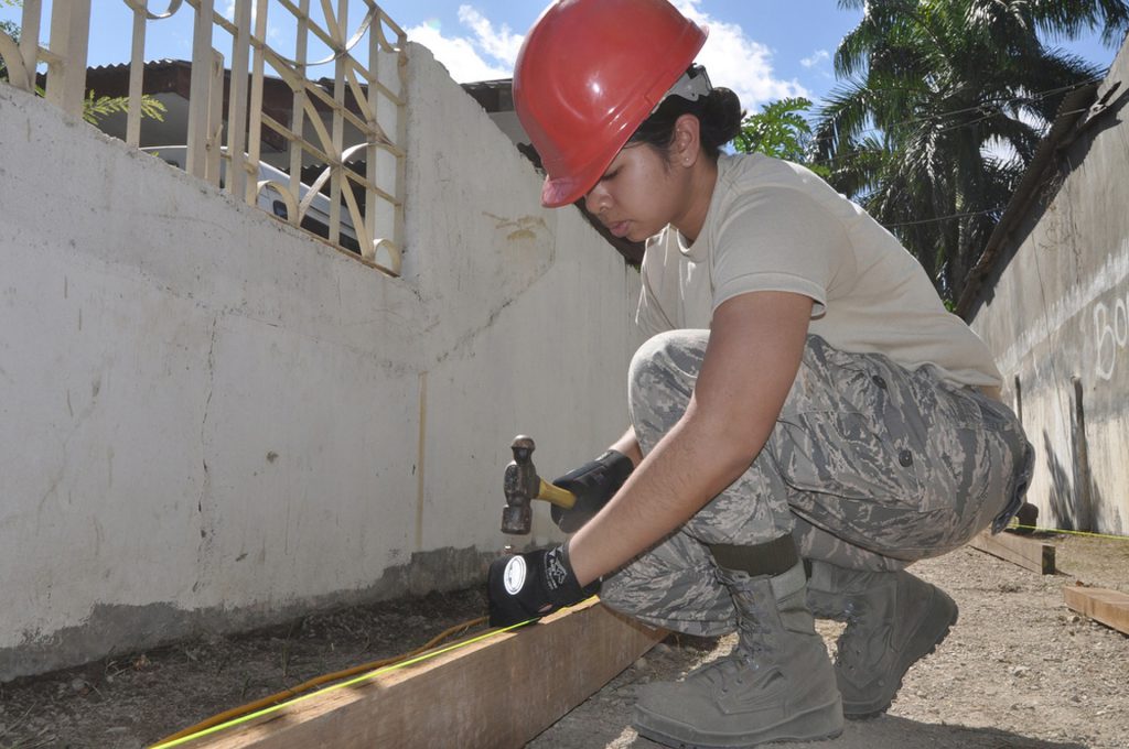 Women in a red hard hammering a nail into a piece of wood on a jobsite. 