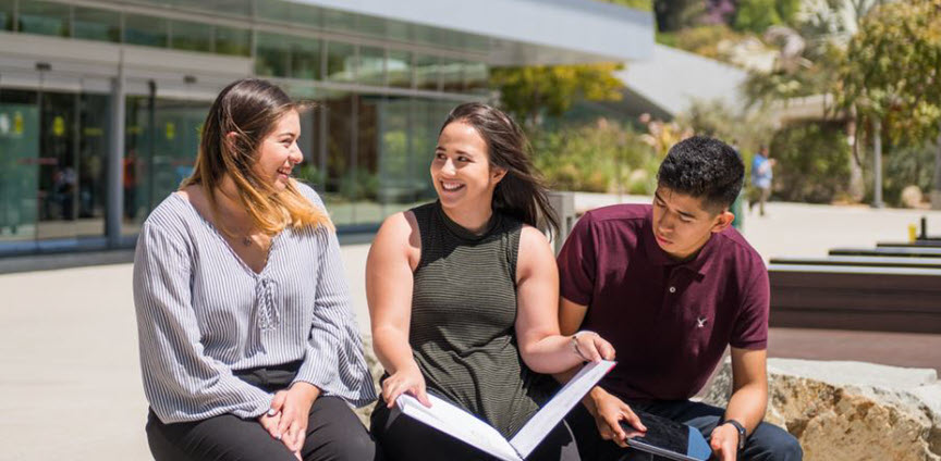 Three students sitting together on campus, one with a textbook open 